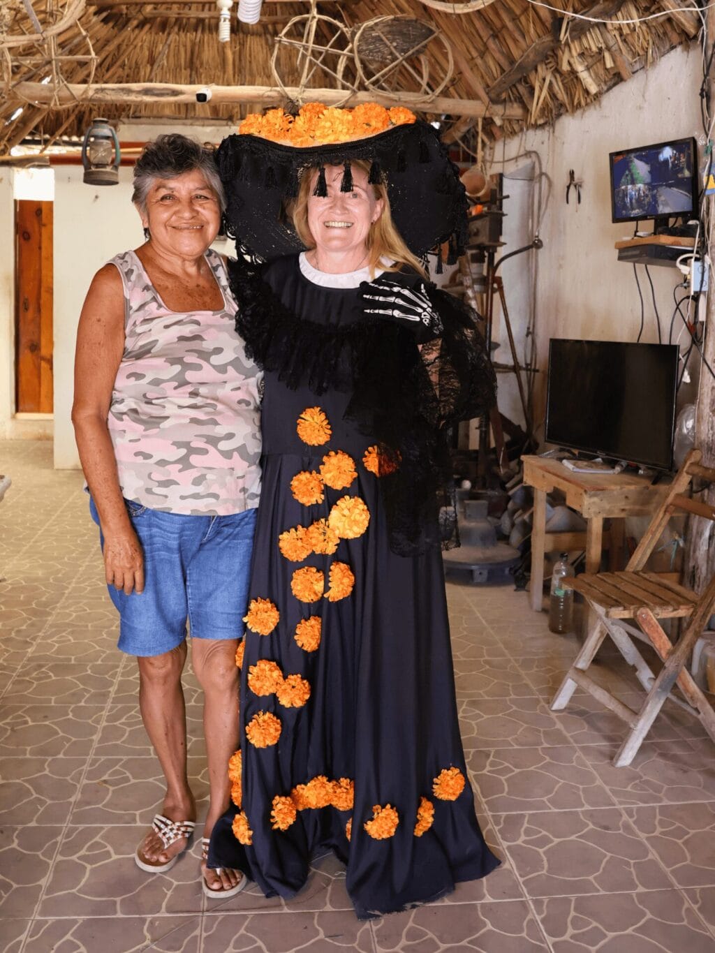 Eine mexikanische Frau mit schwarz-grauen Haaren, blauen kurzen Hosen und einem hellen, gemusterten Träger-T'Shirt steht neben einer blonden Frau, die ein schwarzes Kleid und einen schwarzen Hut mit orangefarbenen Pompoms trägt. Sie befinden sich in einer Halle unter einem Strohdach.